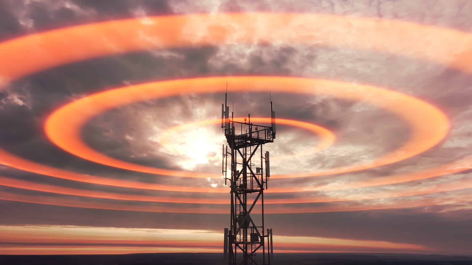 A telecommunications tower stands against a dramatic sky with swirling orange and gray clouds. The tower is equipped with antennas and surrounded by concentric ripples of light, which create a visual effect around it.