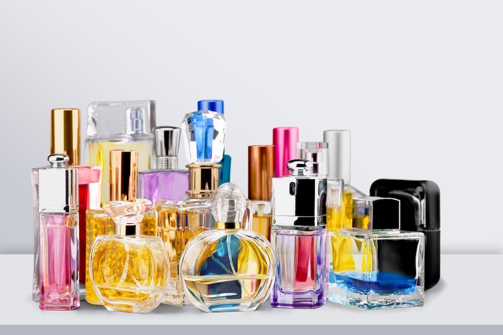 An assortment of perfume bottles of various shapes and colors arranged on a gray surface. The bottles feature glass and metallic designs, with some showcasing colored liquids in clear or tinted glass.