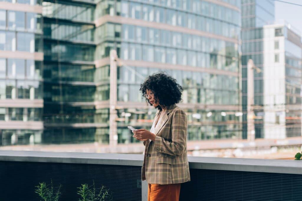 A woman with curly hair wearing a plaid blazer and orange pants is standing outdoors, looking at her smartphone. Modern glass buildings are visible in the background, reflecting sunlight.