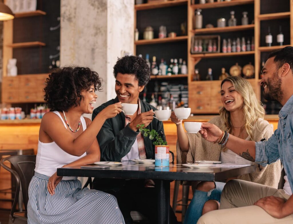 Four friends enjoying coffee at a table in a cozy café. They are smiling and engaging with each other, holding white cups. The background features shelves filled with various food and drink items, creating a warm atmosphere.