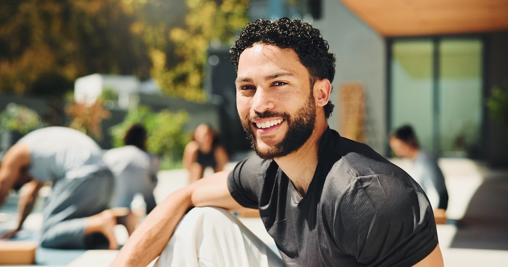 A smiling man with curly hair sits on a yoga mat, wearing a dark shirt and light-colored pants. In the background, several people practice yoga outdoors in a modern setting with greenery and large windows.