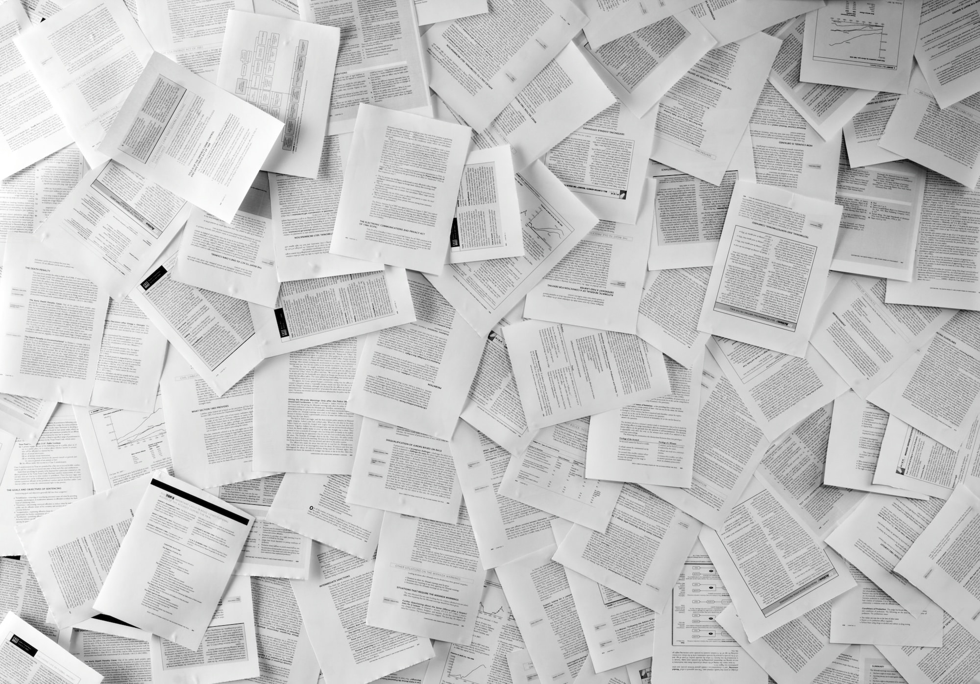 A large number of printed pages scattered across a surface, featuring text and diagrams in black and white. The pages are overlapping, creating a chaotic arrangement that emphasizes a sense of abundance and variety of content.