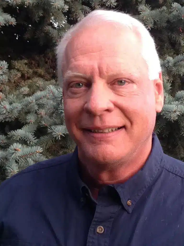 A man with short gray hair and blue eyes is smiling for the camera. He is wearing a dark blue collared shirt. The background features evergreen trees, creating a natural setting.