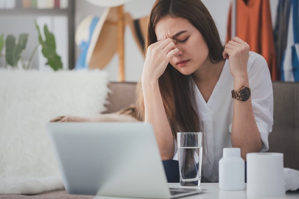 A woman sitting on a couch with her eyes closed and a hand on her forehead, looking stressed. In front of her is a laptop, a glass of water, and two pill bottles. Soft furnishings and colorful clothing hang in the background.