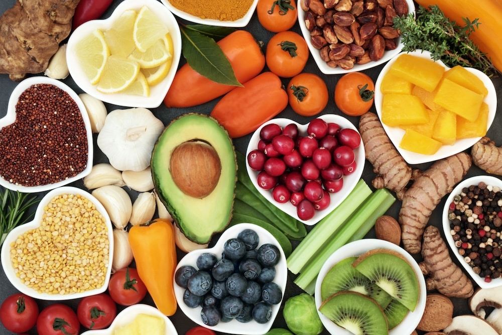 An assortment of colorful fruits, vegetables, and grains arranged in white heart-shaped bowls. Items include avocado, blueberries, tomatoes, yellow bell peppers, mango, nuts, and spices, set on a dark background.