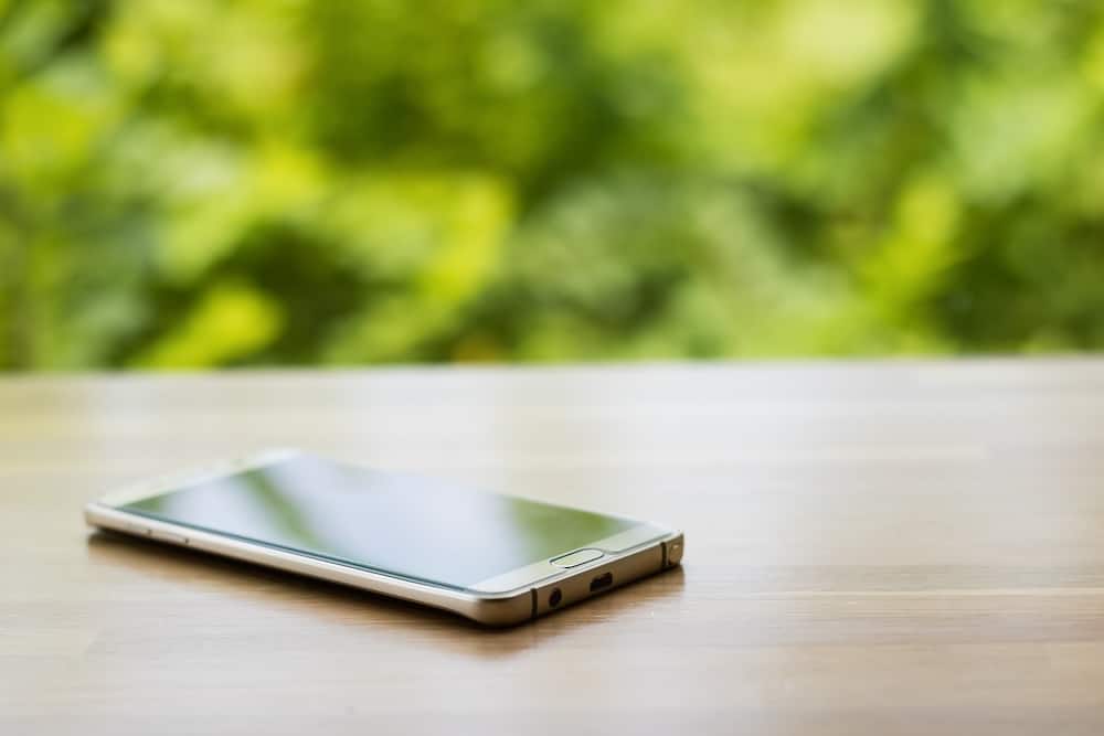 A smartphone lies on a wooden surface, with its screen reflecting light. Background features blurred greenery, creating a natural and serene setting.