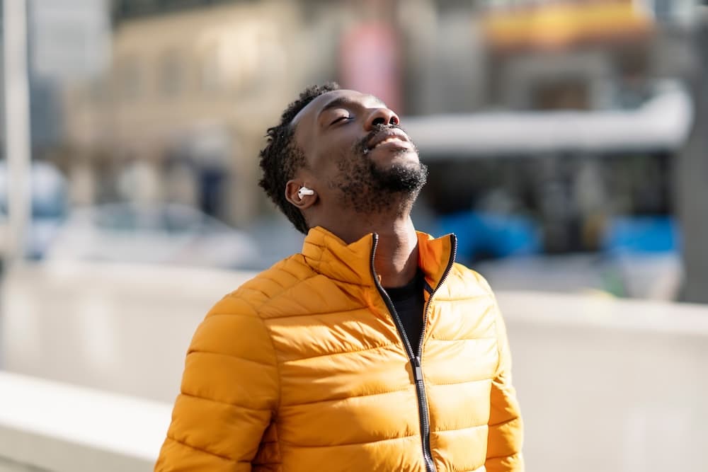 A man wearing an orange puffer jacket with earbuds is standing outdoors, tilting his head back slightly with his eyes closed. Urban buildings and a bus can be seen in the background, indicating a city setting.