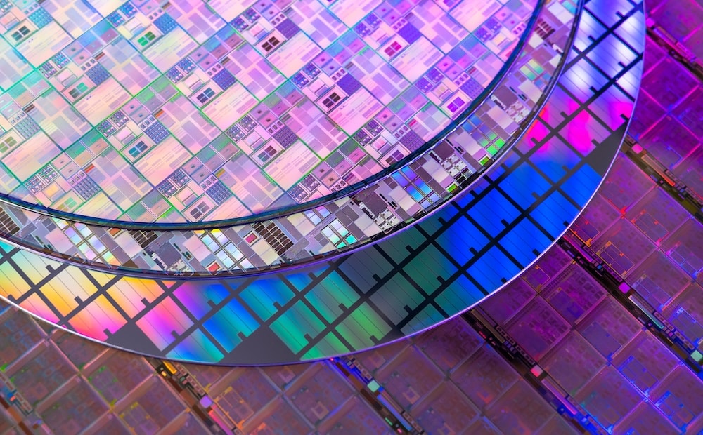 Close-up of semiconductor wafers stacked together, featuring colorful, reflective patterns and a grid of integrated circuits. The wafers display intricate designs, indicative of advanced microelectronic manufacturing.