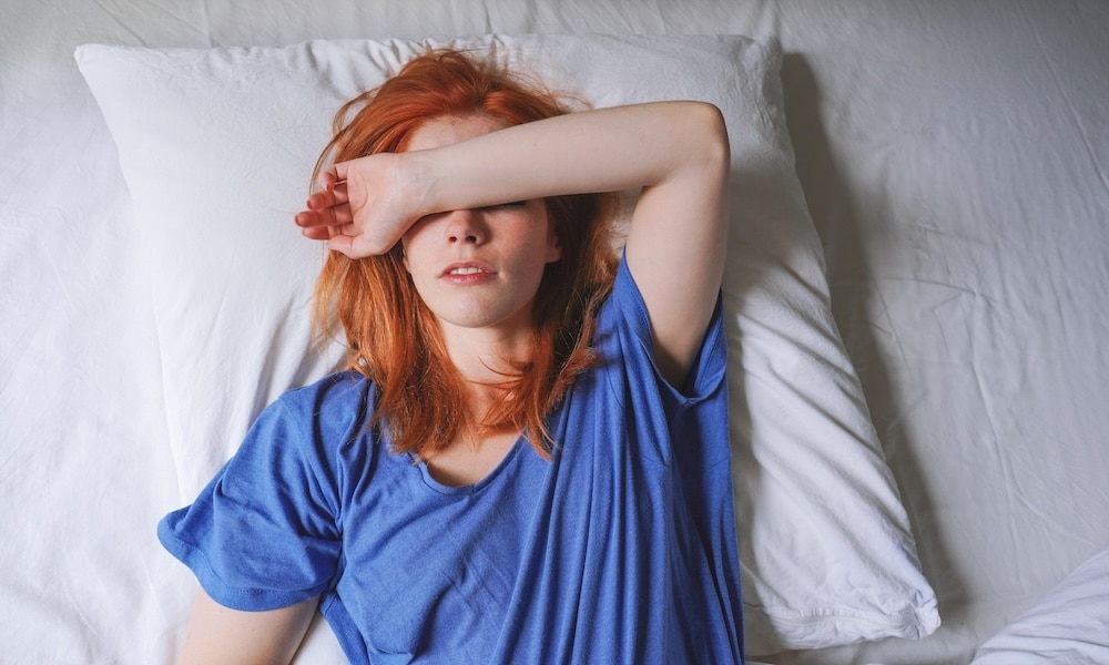 A woman with red hair in a blue t-shirt lies on a bed, resting her arm across her forehead. She appears to be waking up in a bright room, with white bedding and pillows around her.