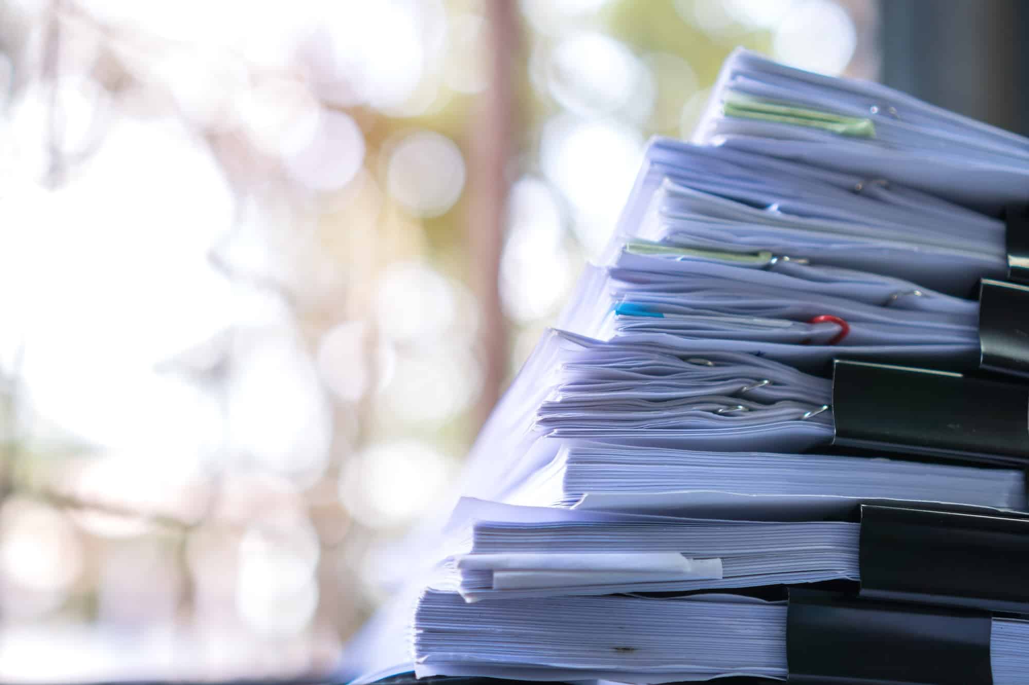 A close-up view of a stack of organized documents secured with black paperclips, with soft blurred background suggesting natural light filtering through, creating a bokeh effect.