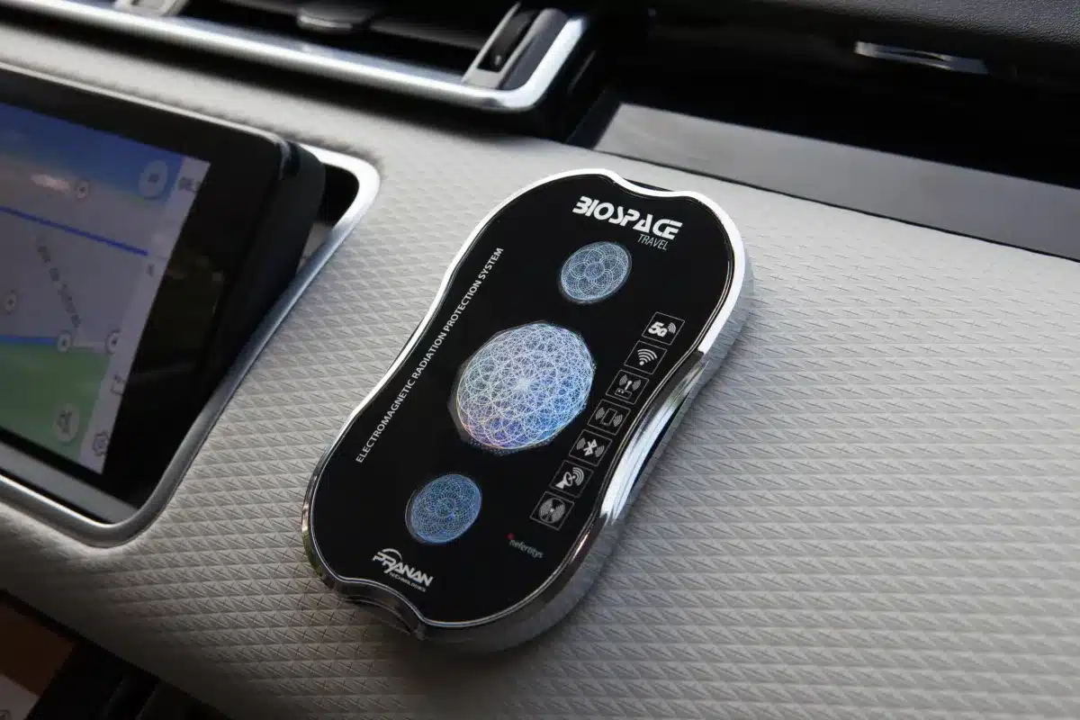 A black and silver electronic device with a touchscreen interface and various buttons, placed on a textured dashboard near a navigation screen in a vehicle. The device shows a circular light display in the center.