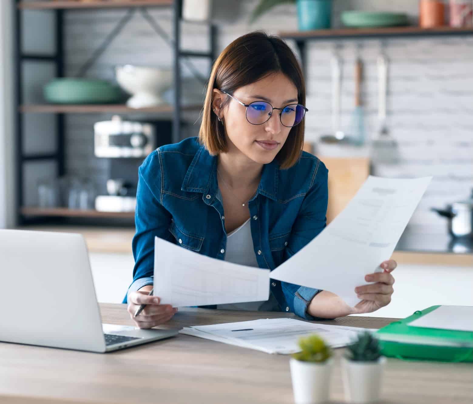 A woman with short brown hair and glasses is sitting at a desk with a laptop open in front of her. She is reviewing documents, holding two sheets of paper in one hand. The background features a modern kitchen setting with shelves and plants.