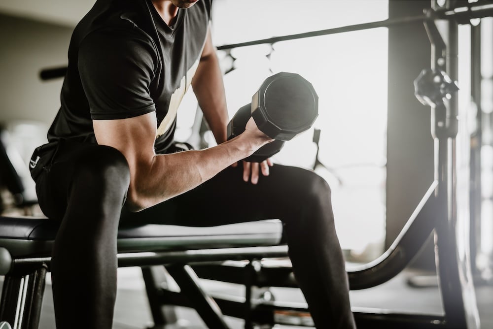 A person sitting on a weight bench in a gym, performing a bicep curl with a dumbbell. They are wearing a black athletic shirt and leggings, with gym equipment visible in the background.